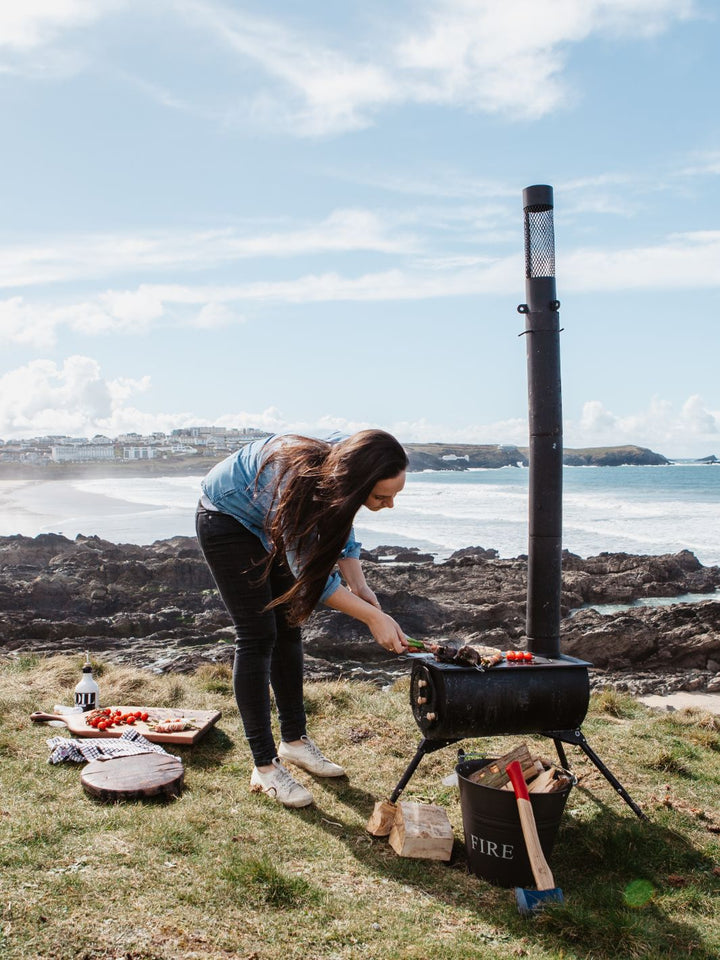 Anevay Frontier Plus - set up on the Cornish coast with a woman bent over the stove, cooking lunch - Outdoor Wood Burning Stove